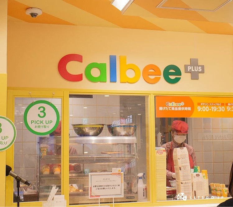 Calbee+ 專賣店櫃檯,上方掛有彩色品牌標誌,並設有領餐區指示牌。